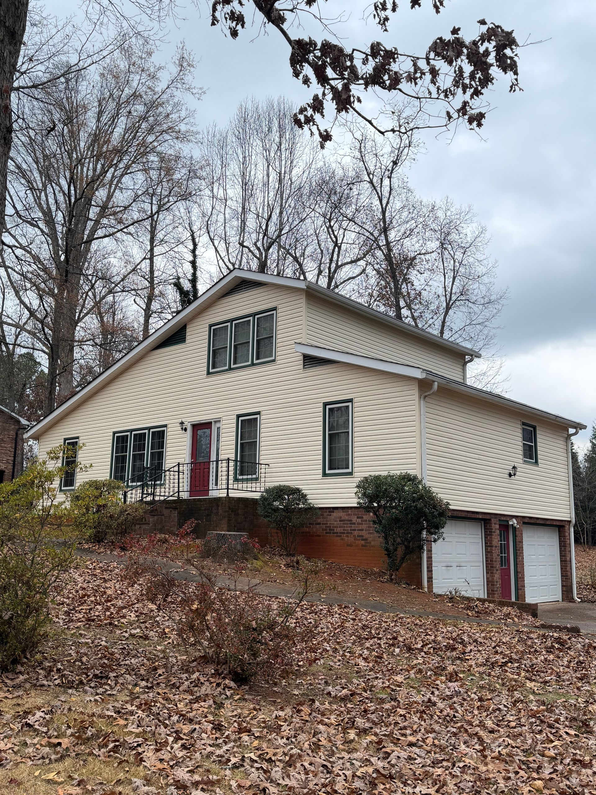 A two-story, cream-sided house with a garage underneath, set on a sloped, leaf-covered lot surrounded by trees.