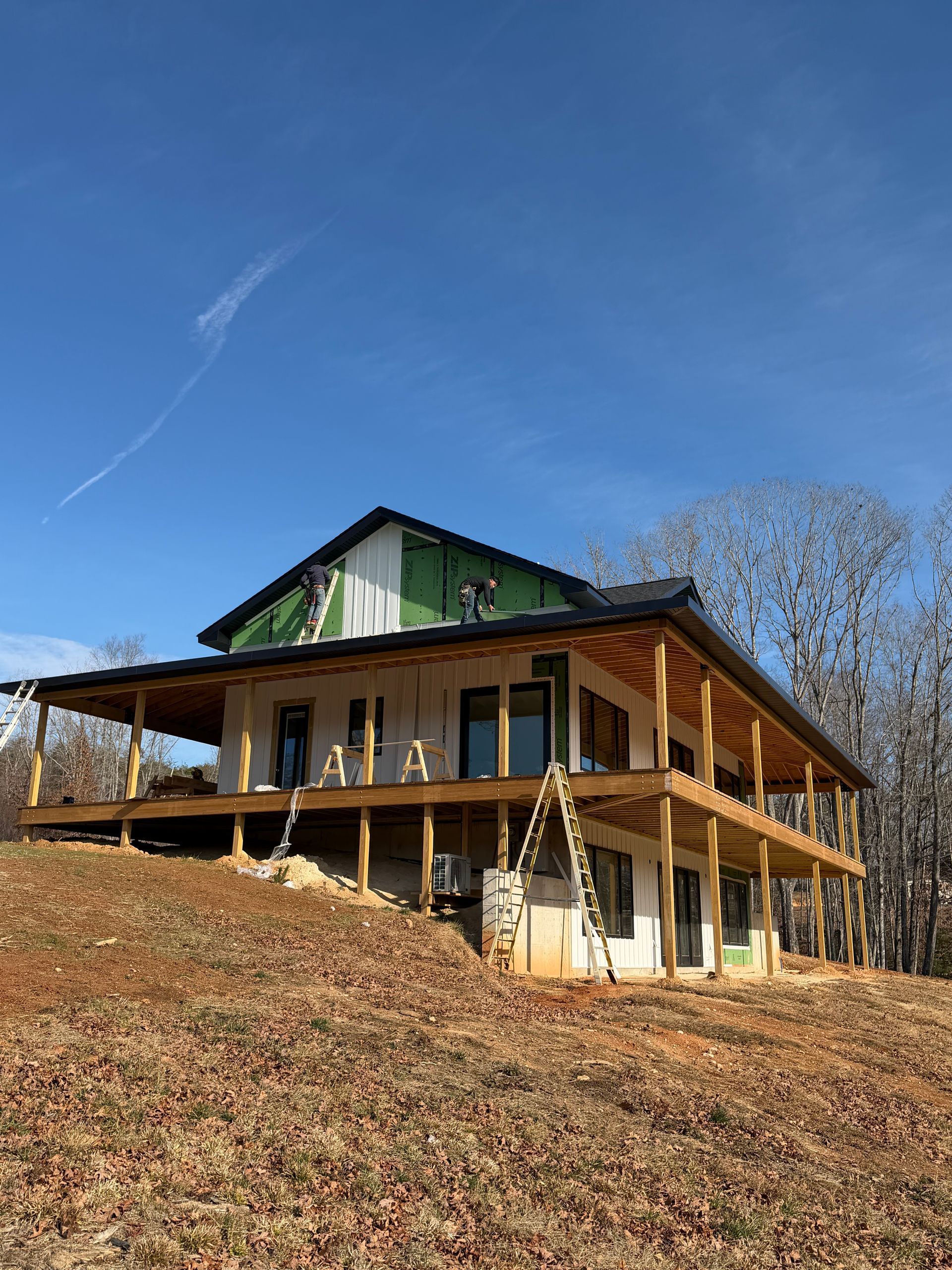 A house under construction with a wraparound porch stands on a grassy, leaf-covered hill under a clear blue sky.