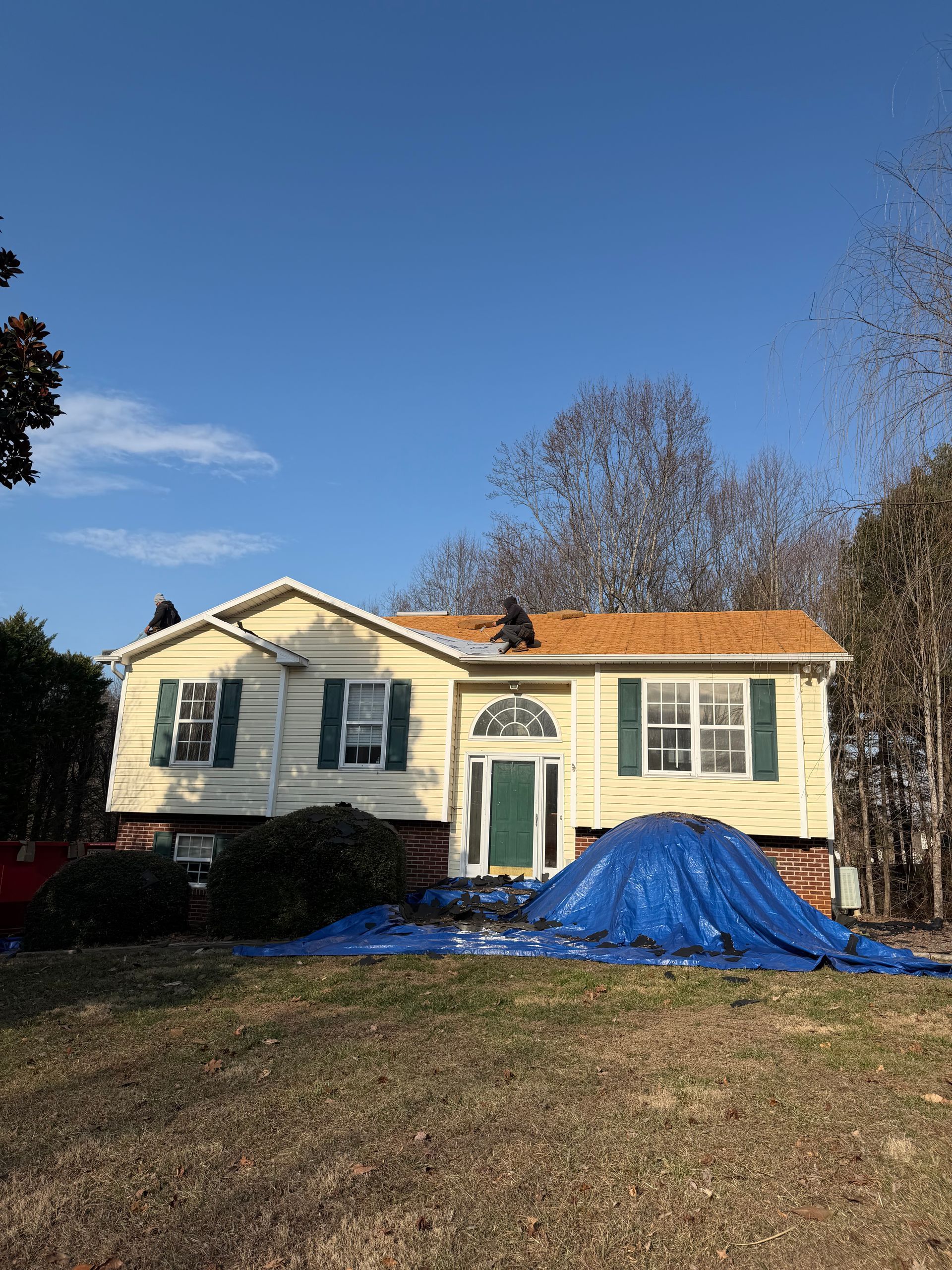 A yellow two-story house with a partially replaced wood roof and a large blue tarp covering the front yard and foundation.