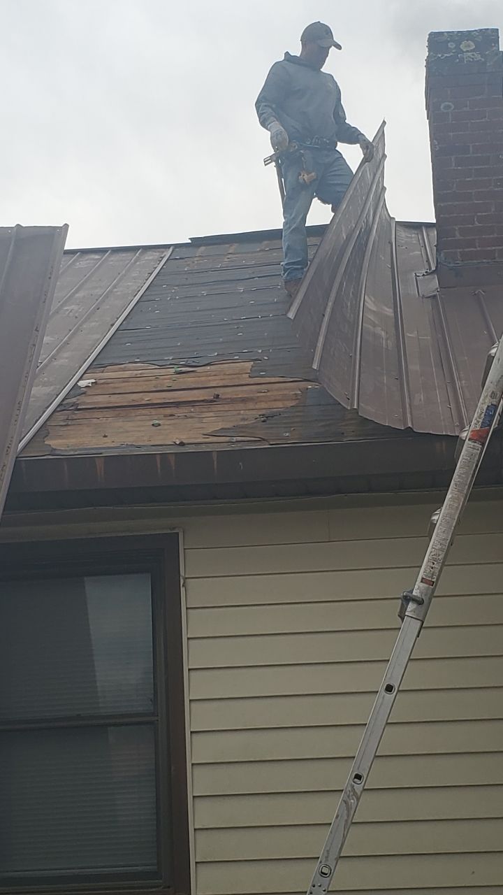 A person in work clothes on a house roof peeling back old metal roofing to reveal damaged wooden decking below.