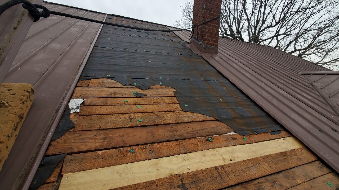 An unfinished roof section showing exposed wooden planks, black underlayment, and metal roofing panels near a brick chimney.