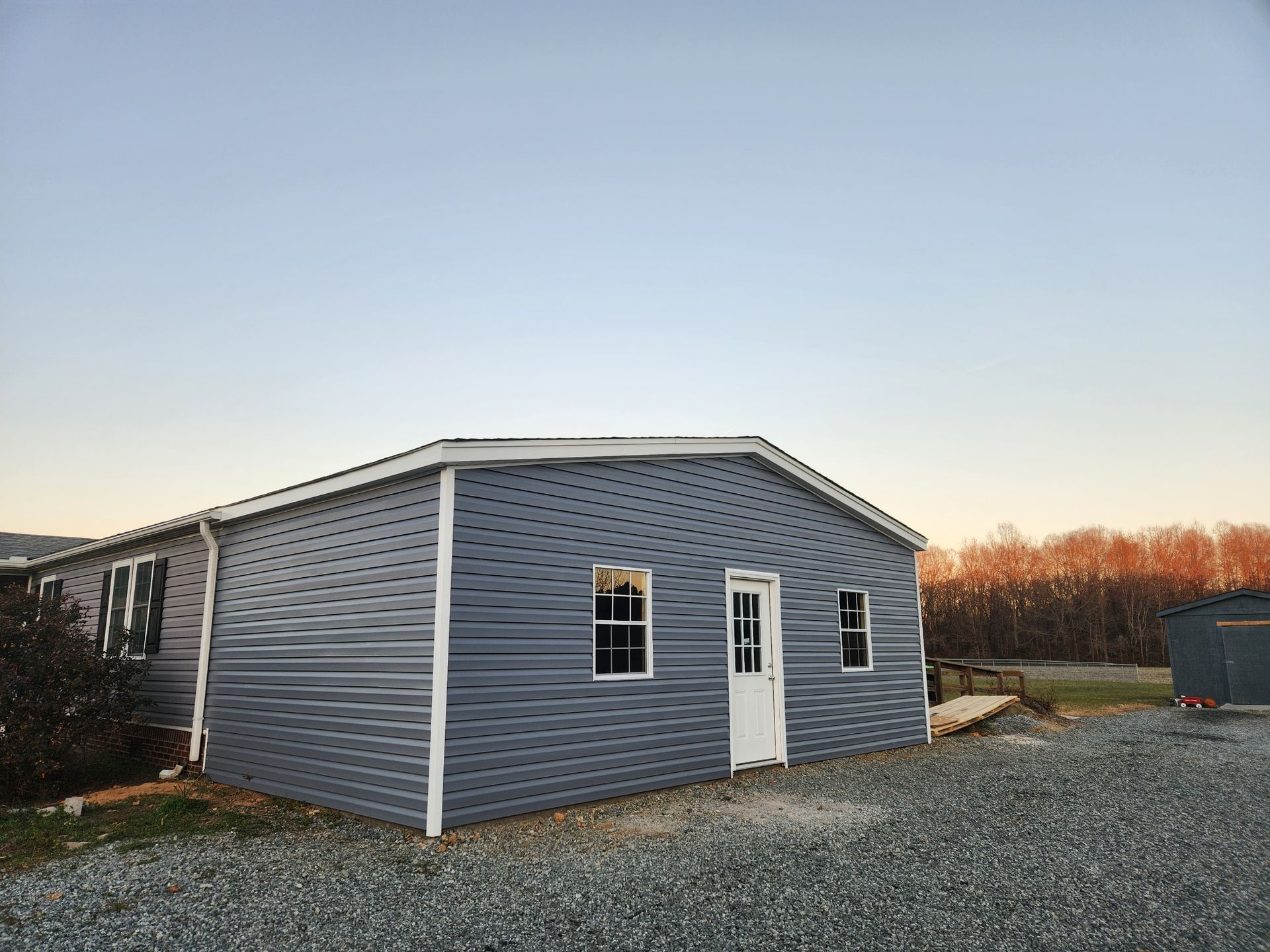 A gray-sided building with a white door and windows sits on a gravel lot under a clear, sunset sky.
