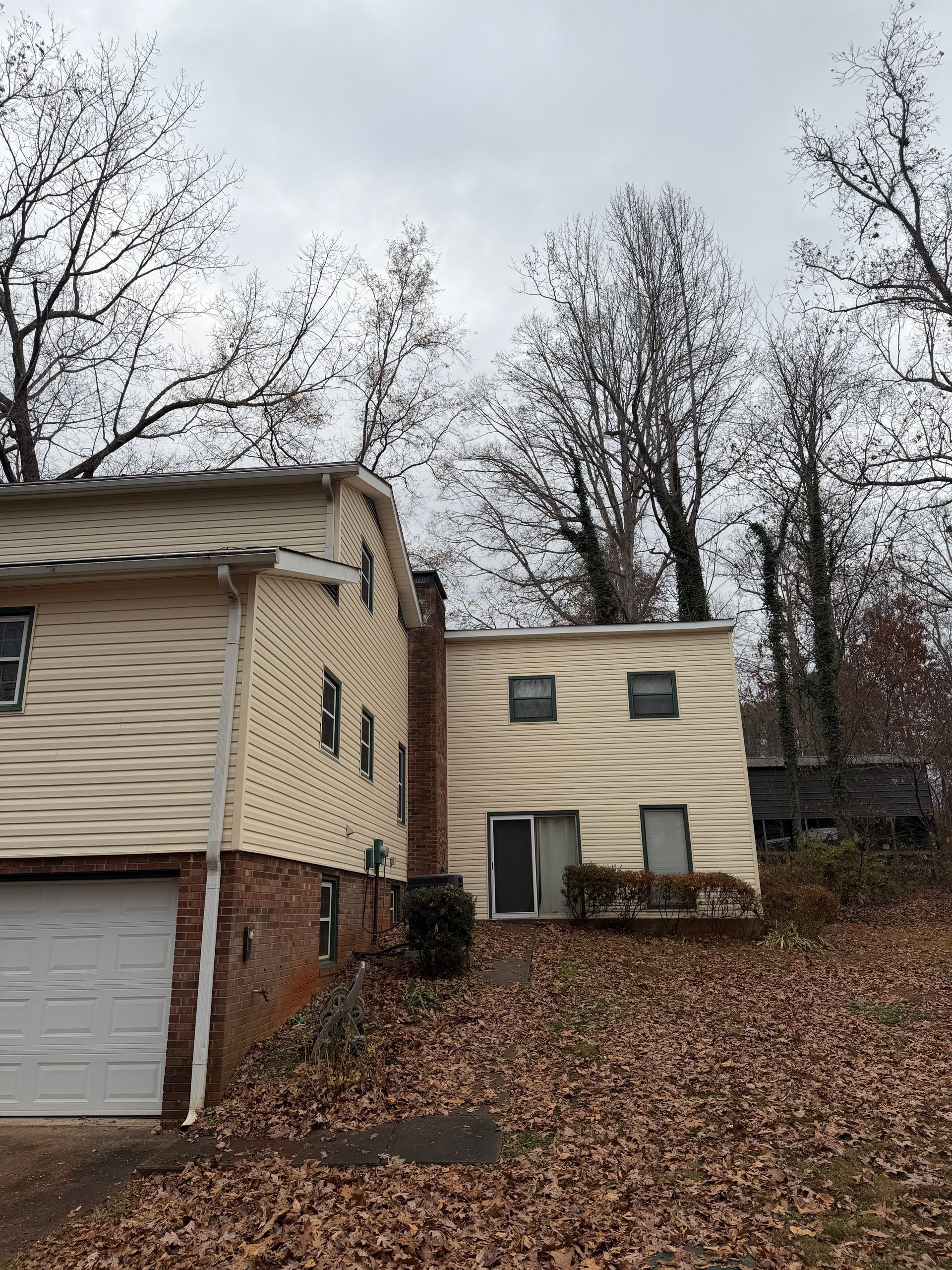 A two-story house with beige siding and a brick base, featuring a garage and surrounded by trees covered in fallen leaves.
