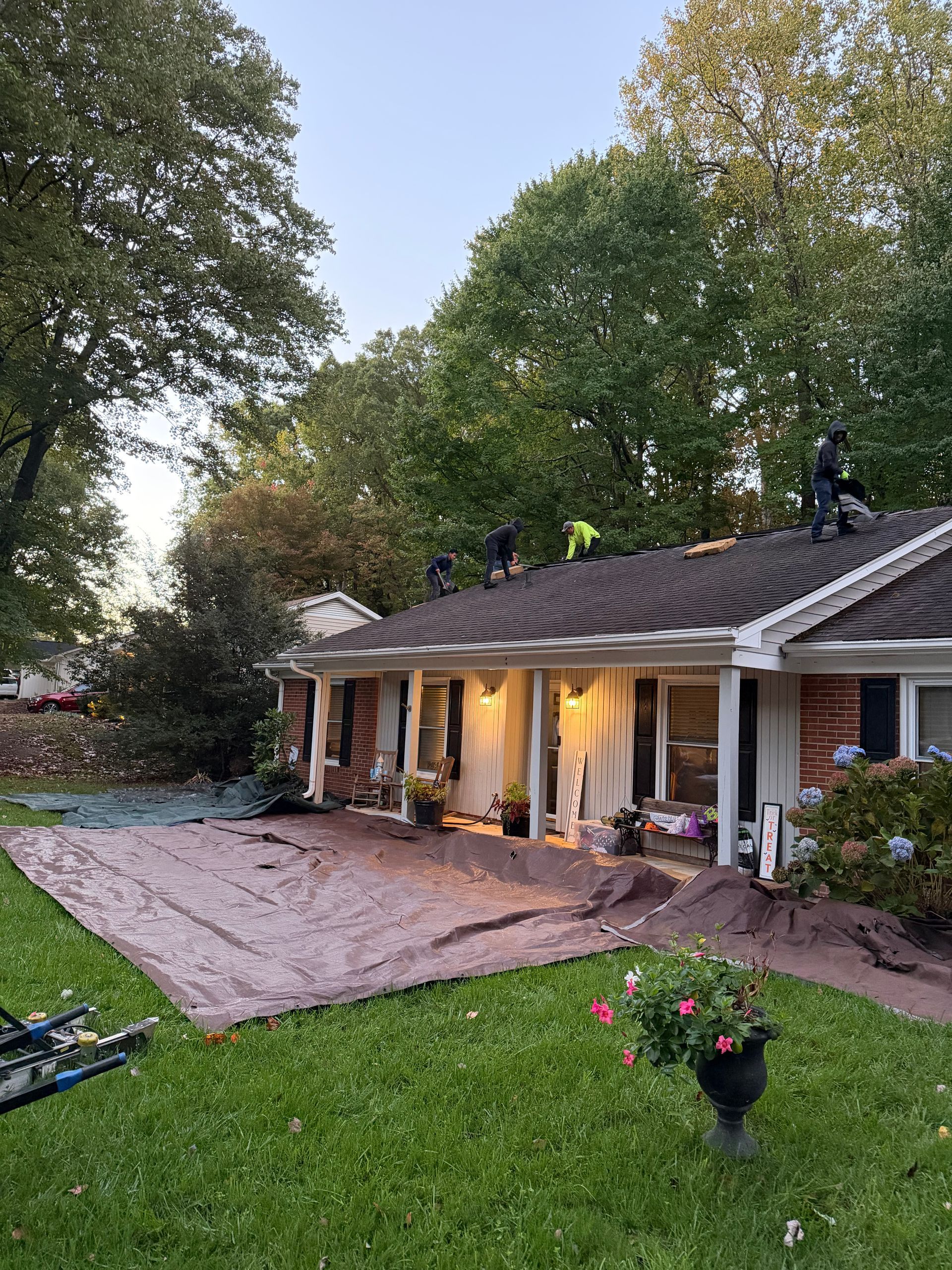 Four roofers work on the shingled roof of a brick house while protective tarps cover the lawn below.