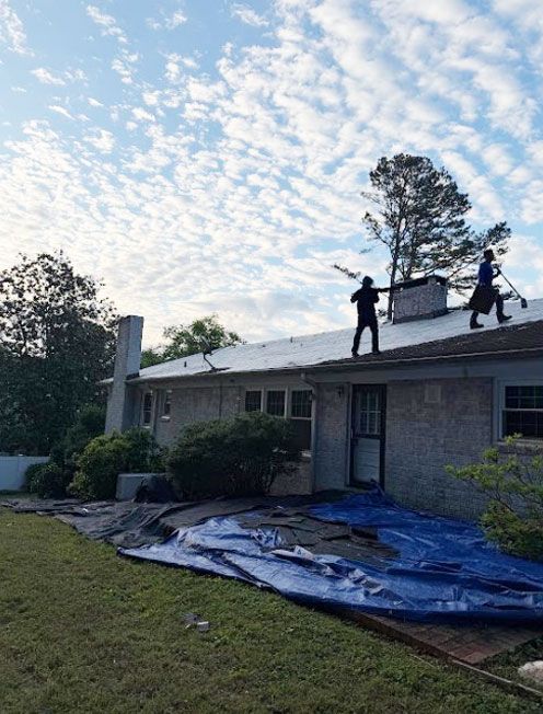 Two workers on a roof repairing storm damage; blue tarp covers the lawn beside a brick house.