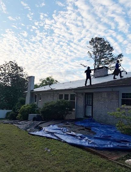 Two workers on a roof repairing storm damage; blue tarp covers the lawn beside a brick house.