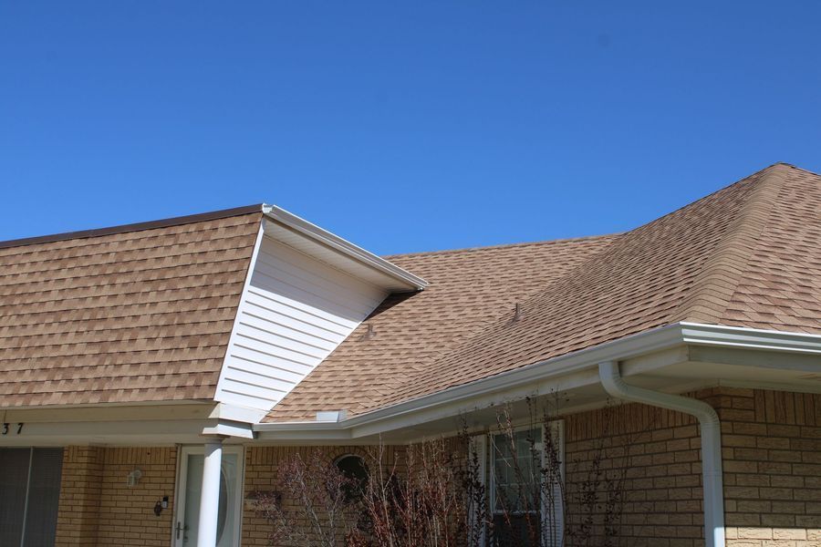 Brown shingle roof with white trim and gutters against a blue sky, on a brick house.