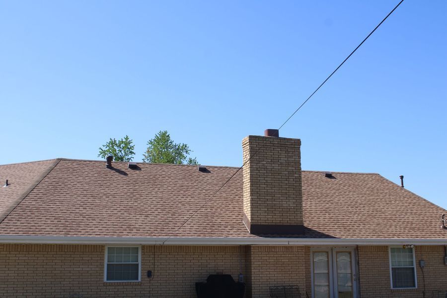 Tan brick house with a brown shingle roof, chimney, and blue sky.