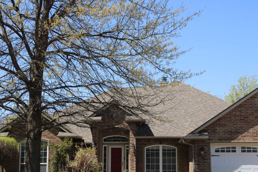 Brick house with brown roof and red door, tree in foreground, bright blue sky.