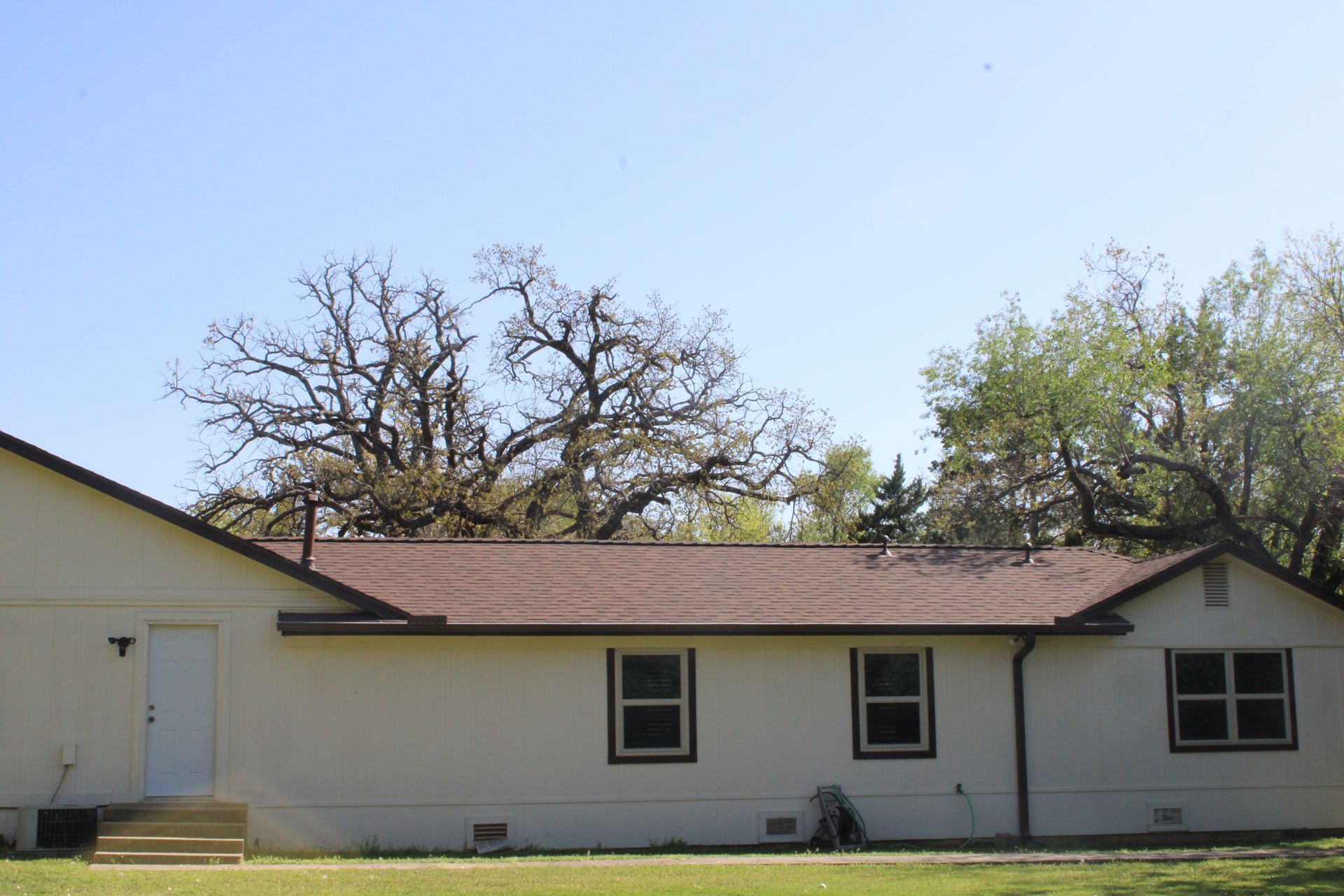 Beige house with a brown roof and green grass, trees in the background, blue sky.