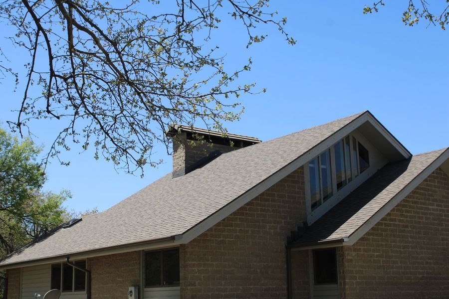 Tan brick house with a brown shingled roof, chimney, and angled window under a blue sky.