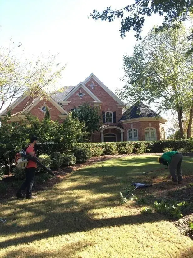 Workers landscaping a brick house yard under trees in bright sunlight