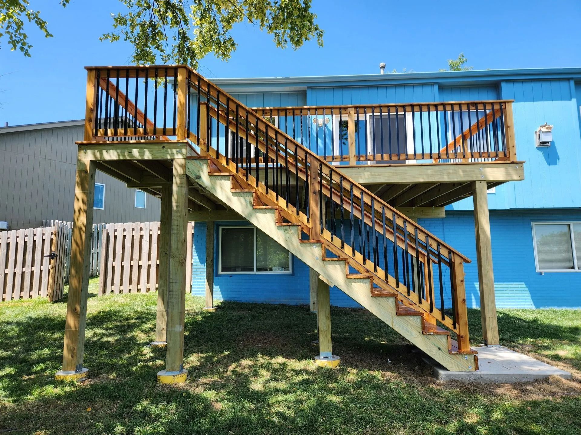 Wooden deck with stairs attached to a blue house, surrounded by green grass and a fence.