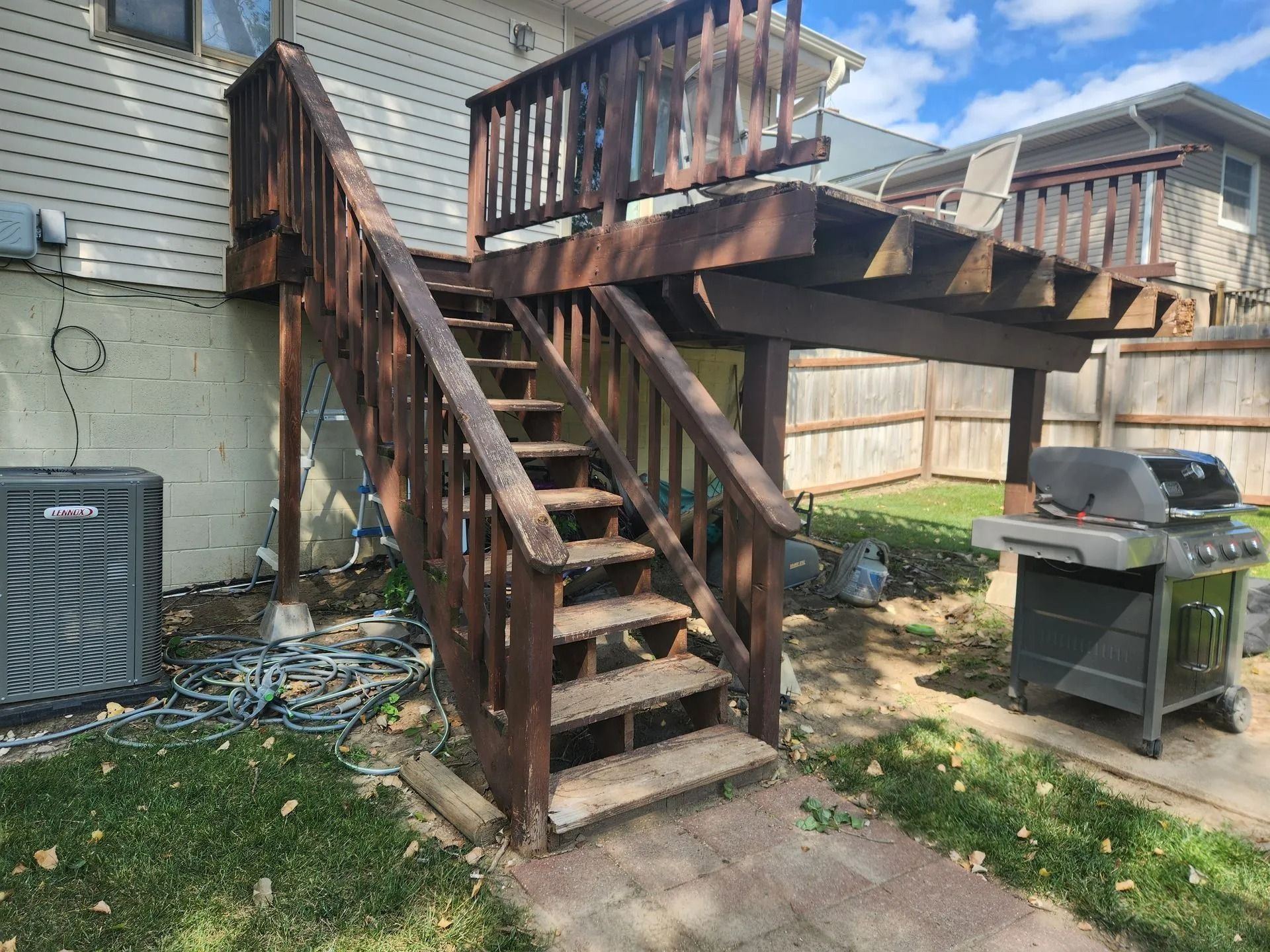 Wooden deck and stairs leading up to it, next to a house with a grill below.