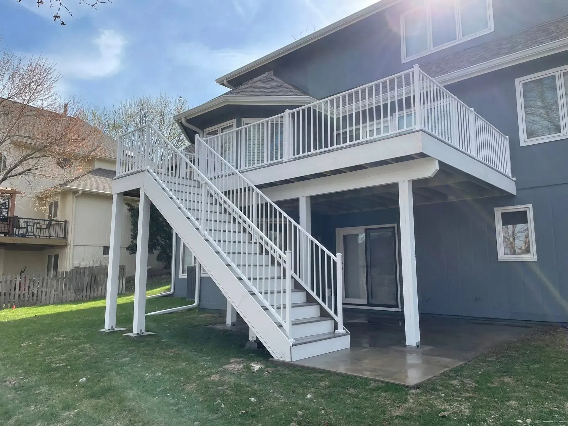 A two-story deck with white railing and stairs against a gray house, on a grassy lawn.