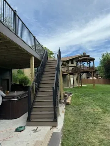 Wooden staircase leads from deck to yard. Black railing with a hot tub visible on the left, gazebo in the background.