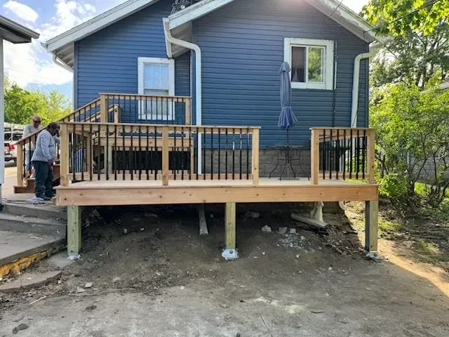 New deck attached to a blue house. Two people working. Wooden deck with black railings.