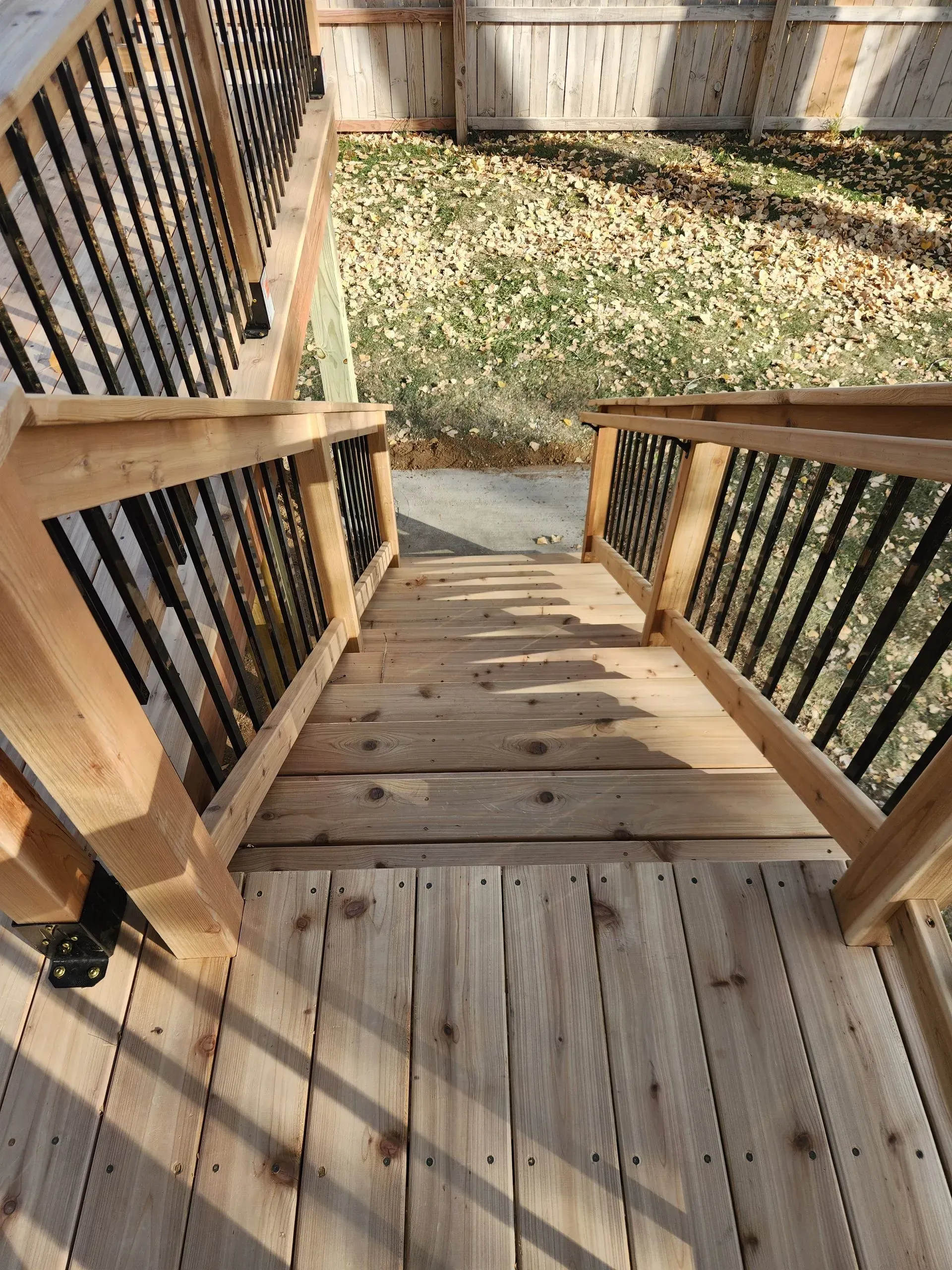Wooden deck stairs with black railings leading to a concrete path and yard with fallen leaves.