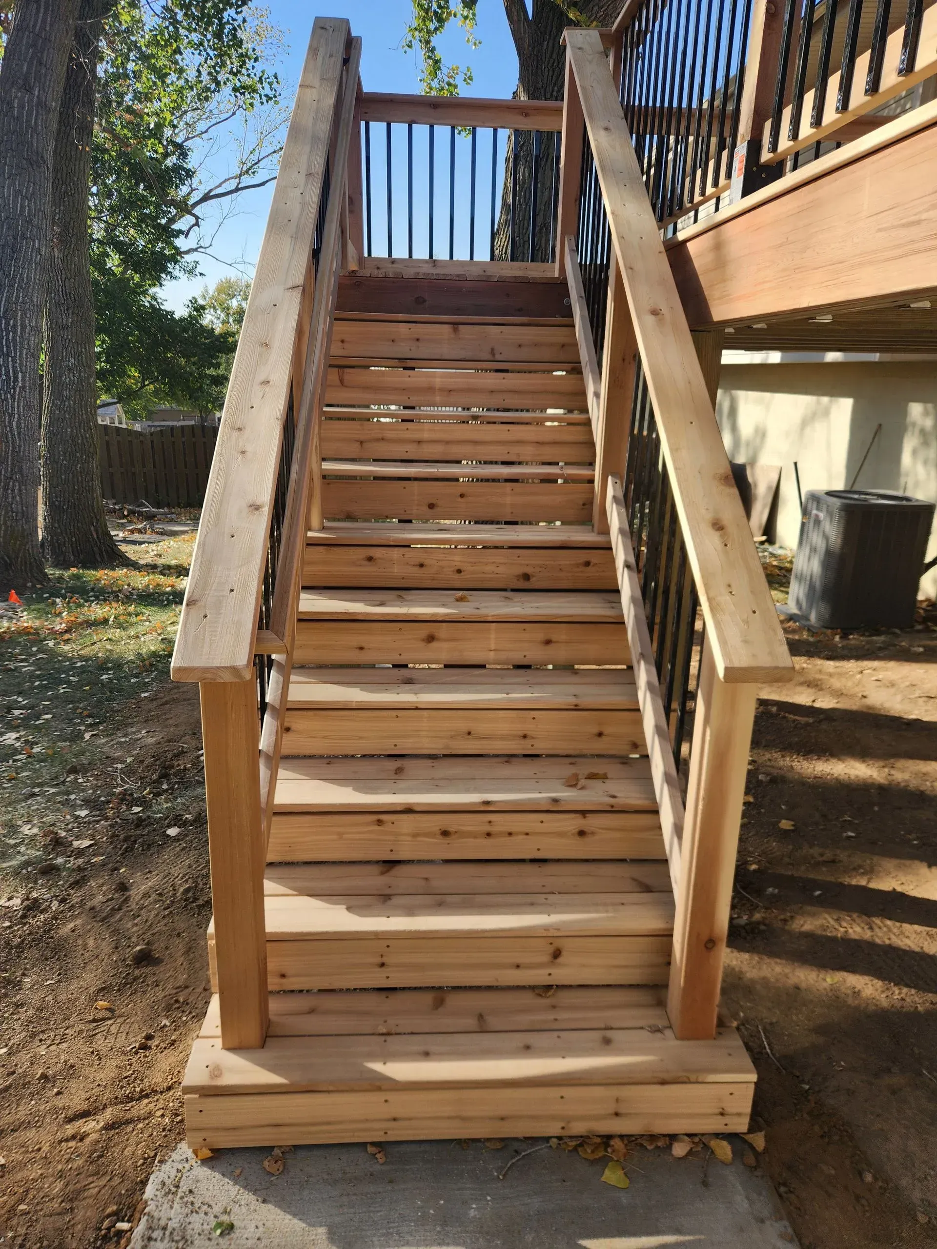 Wooden outdoor staircase leading up to a deck, set against a backdrop of trees and a clear sky.