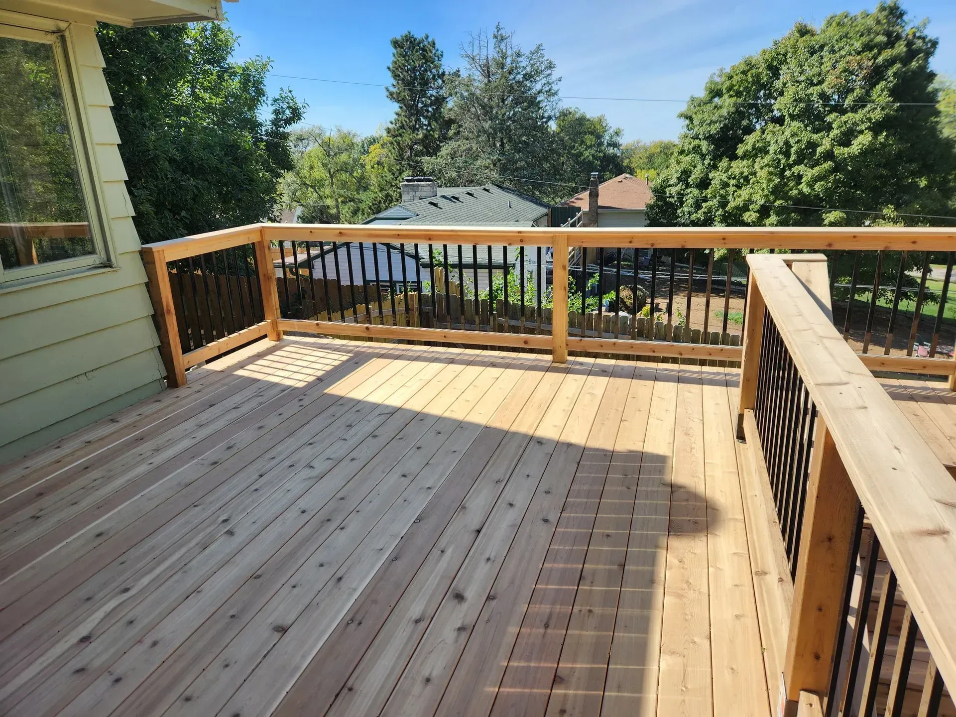 Wooden deck with black railings and light brown boards, overlooking a residential area with trees.