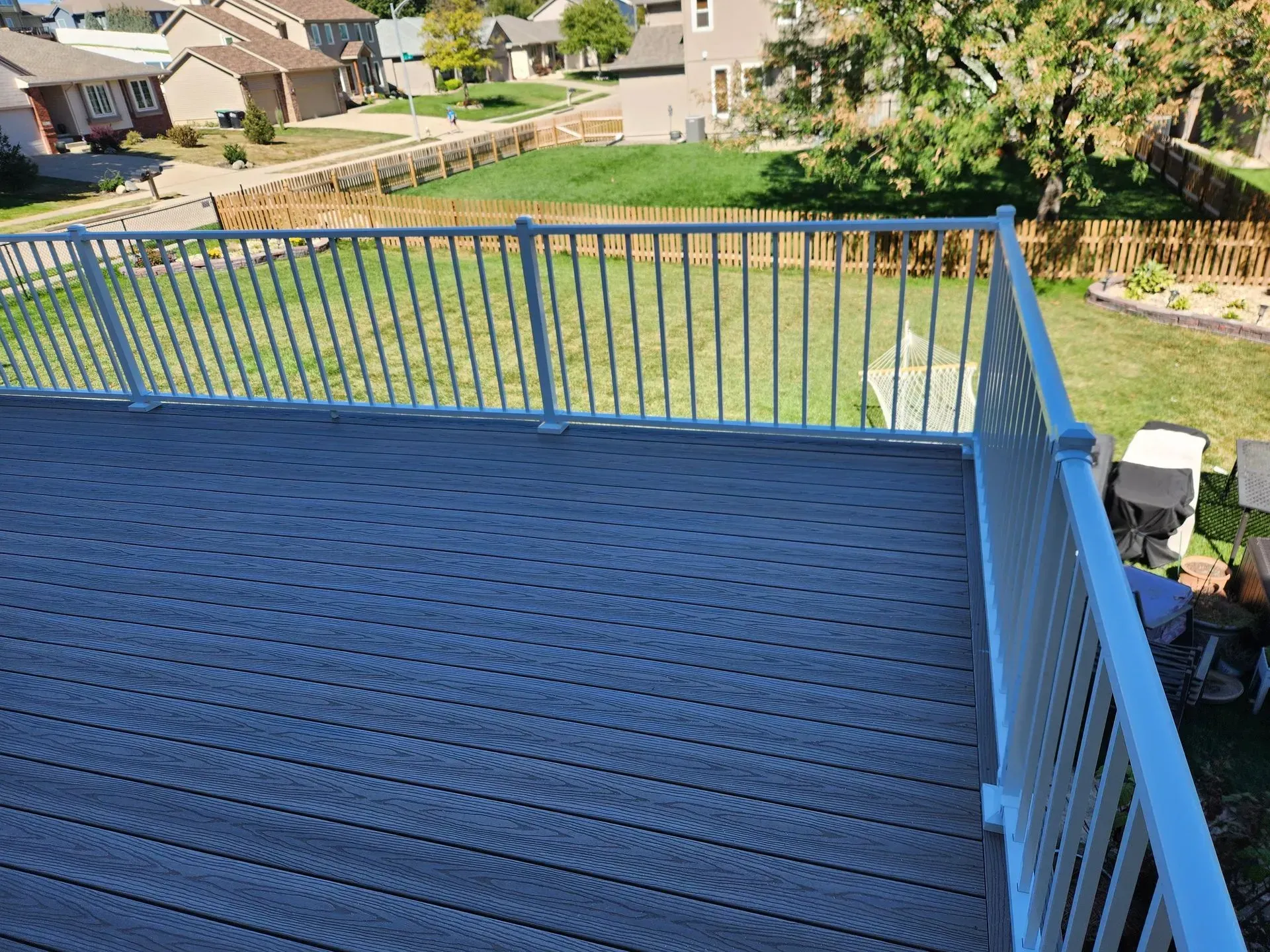 A gray deck with white railing overlooks a green yard and neighborhood.