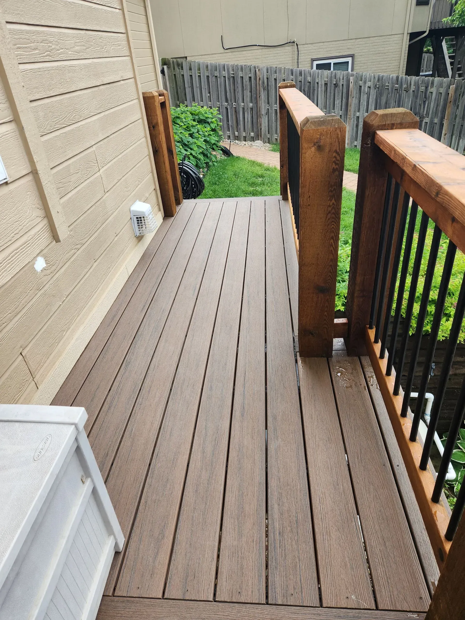 A wooden deck with railing leads to a grassy area. Brown deck boards and railings, with black spindles. Beige siding on the left.