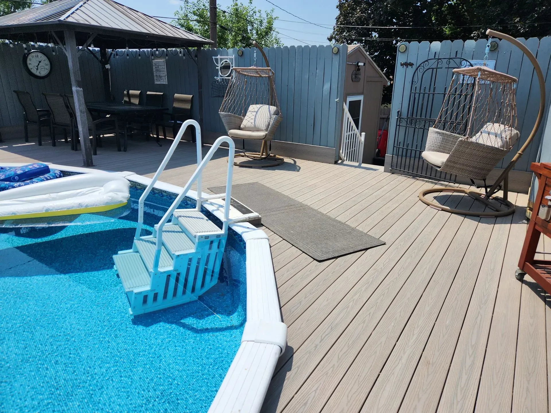 Pool with steps, deck, swings, table, and blue fence on a sunny day.