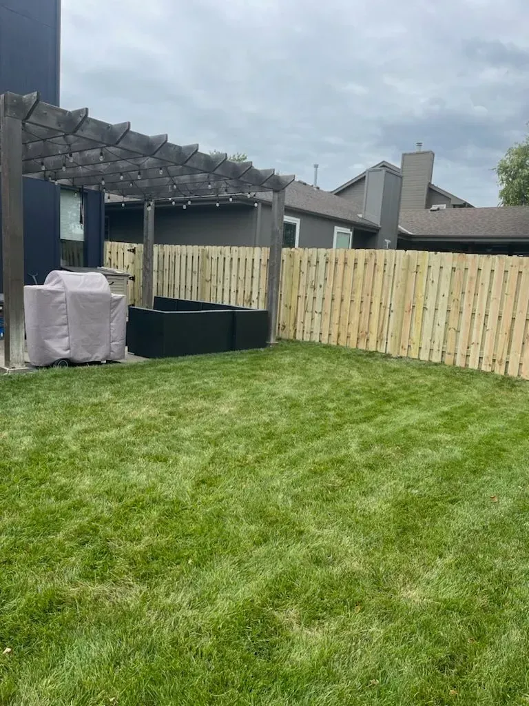 A backyard with green grass, a wooden fence, pergola, and covered grill under a cloudy sky.