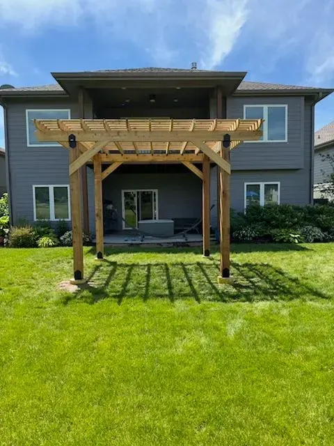 Wooden pergola in a grassy backyard, casting shadows on the lawn. A gray house is in the background.