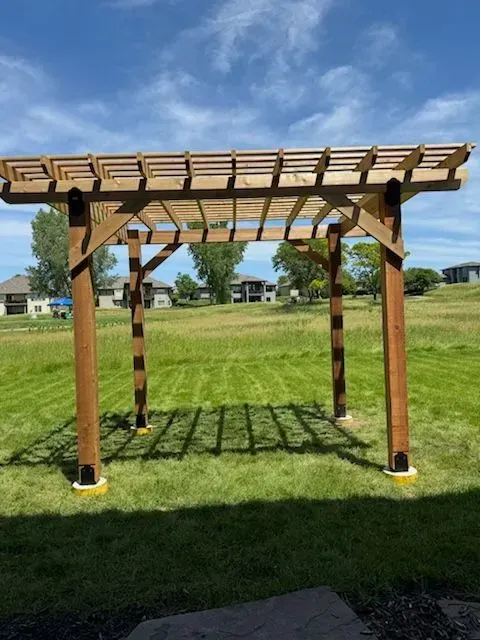 Wooden pergola in a grassy field on a sunny day. Shadows from the structure fall on the ground.