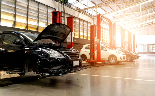 Cars in an auto repair shop, with one black car raised and its hood open in bright sunlight.