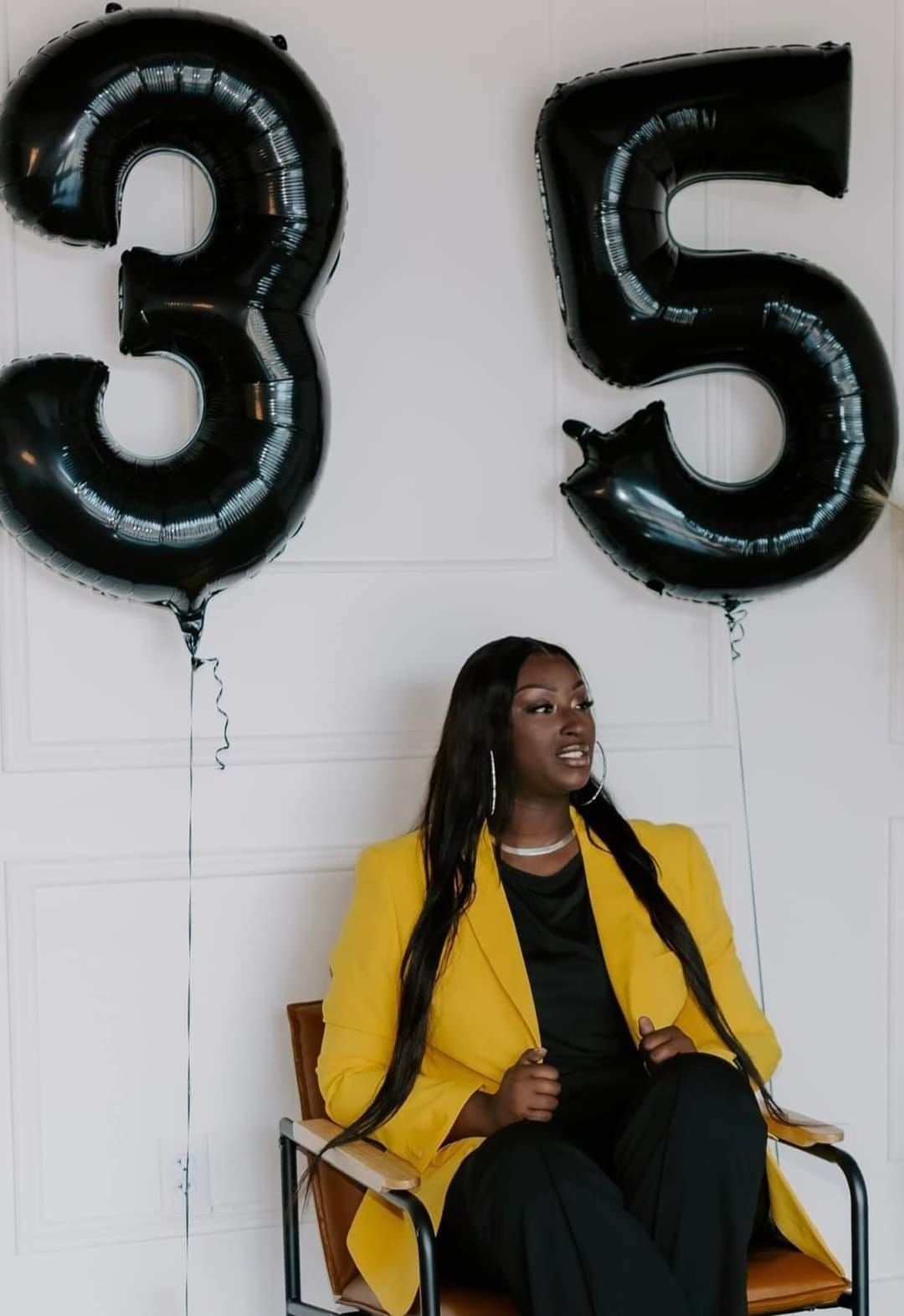 A woman in a yellow jacket is sitting in a chair with black balloons in the shape of the number 35.