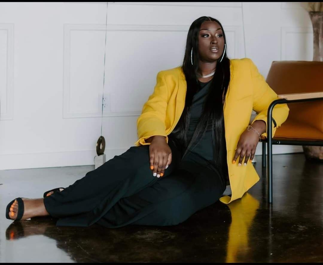 A woman in a yellow jacket sits on the floor next to a chair.