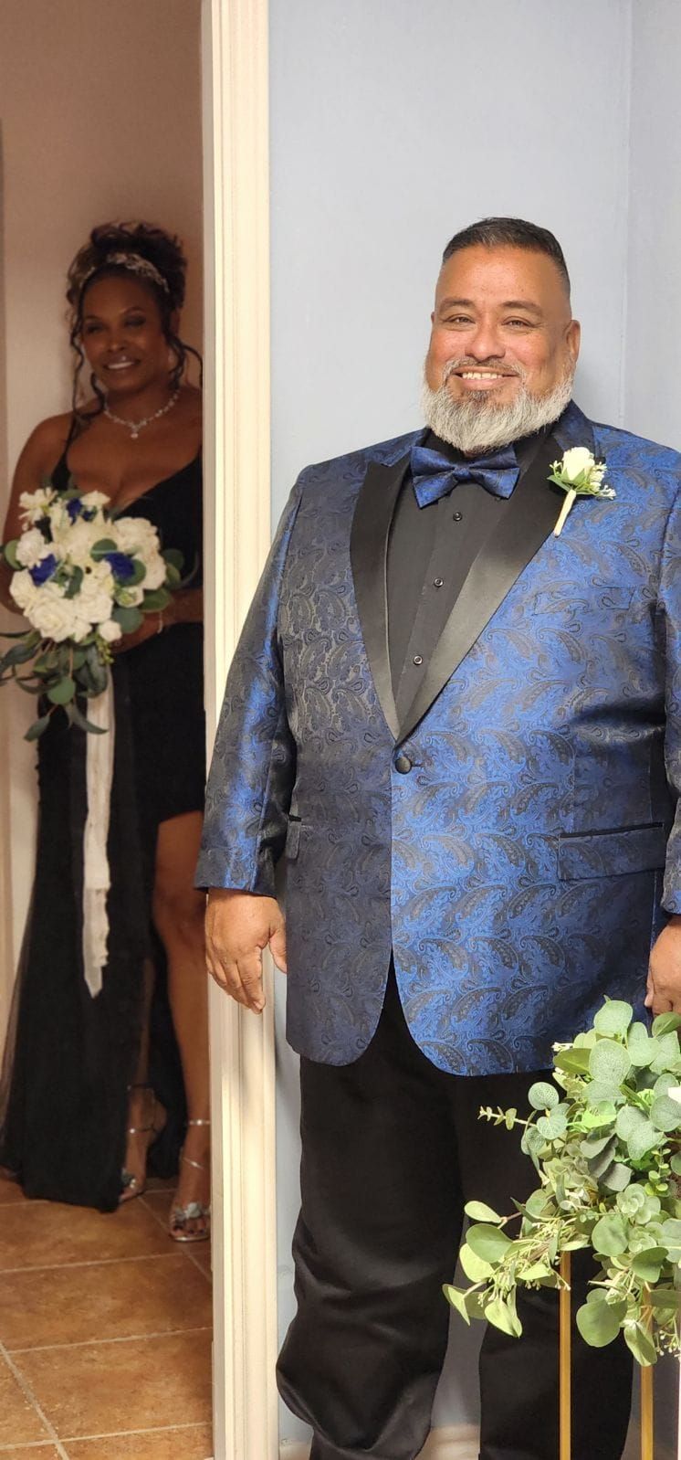 A man in a blue tuxedo stands next to a woman in a black dress holding a bouquet.