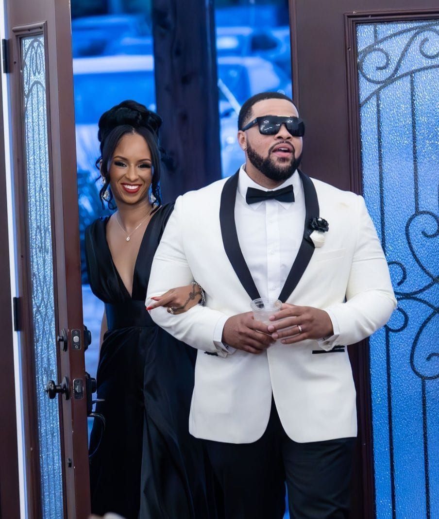 A man in a white tuxedo stands next to a woman in a black dress.