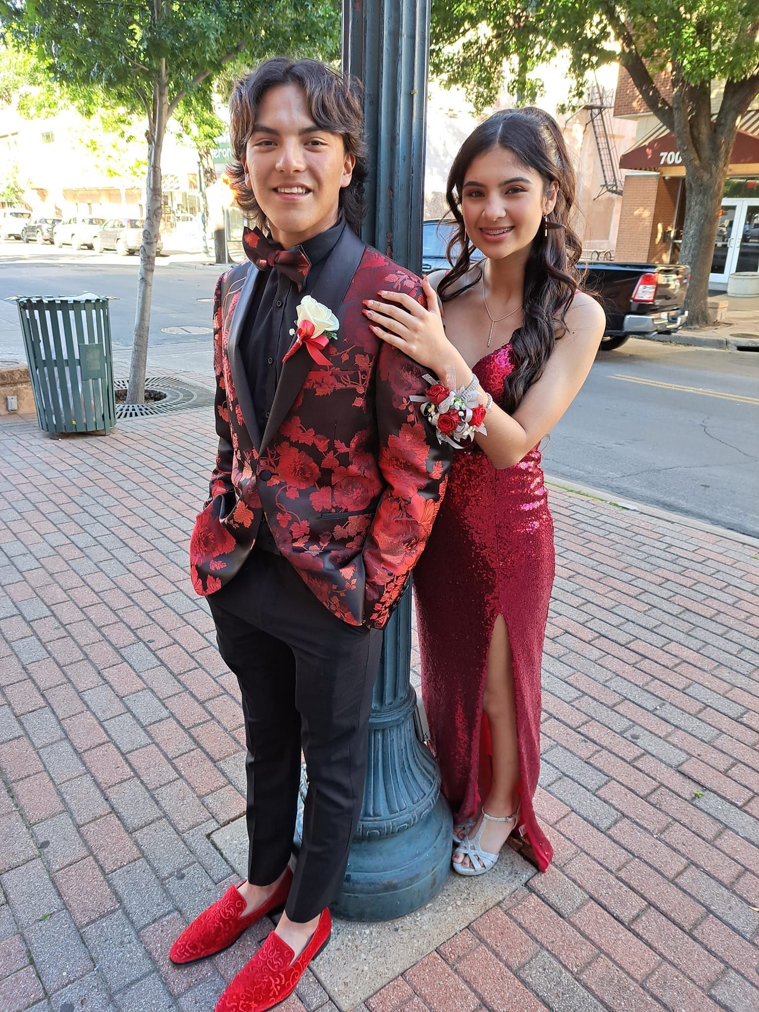 A man in a suit and a woman in a red dress are standing next to each other on a brick sidewalk.