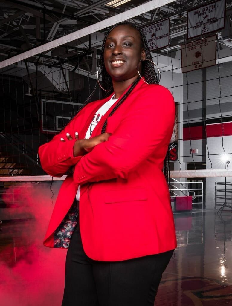 A woman in a red jacket is standing in a gym with her arms crossed.