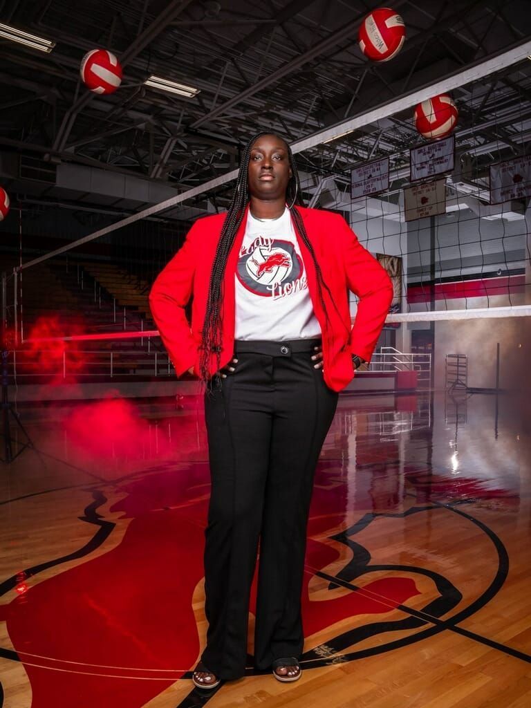 A woman in a red jacket and black pants is standing on a volleyball court.