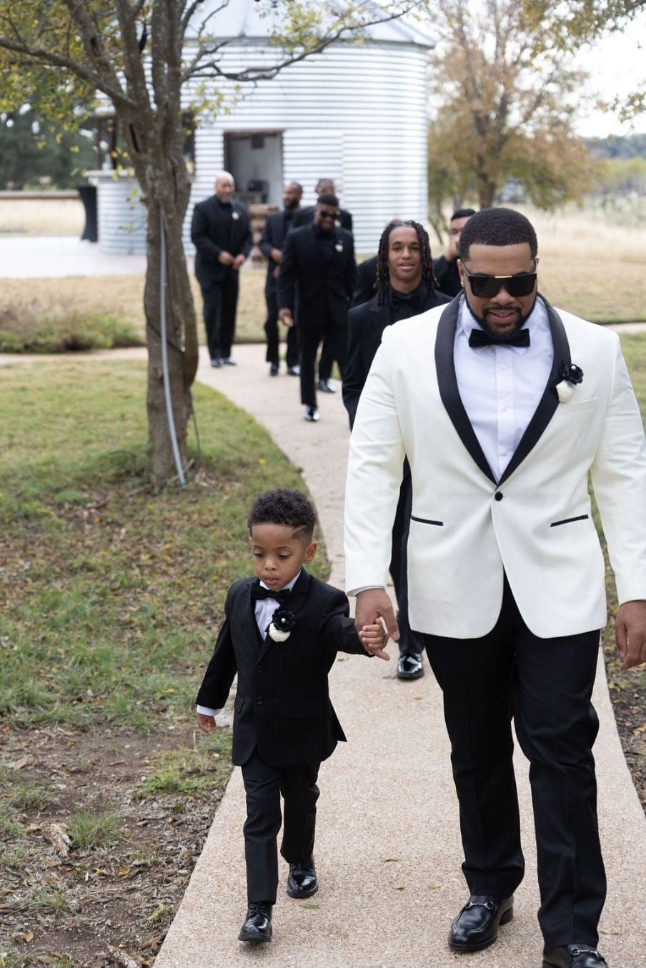A man in a white tuxedo is holding the hand of a little boy in a black tuxedo.