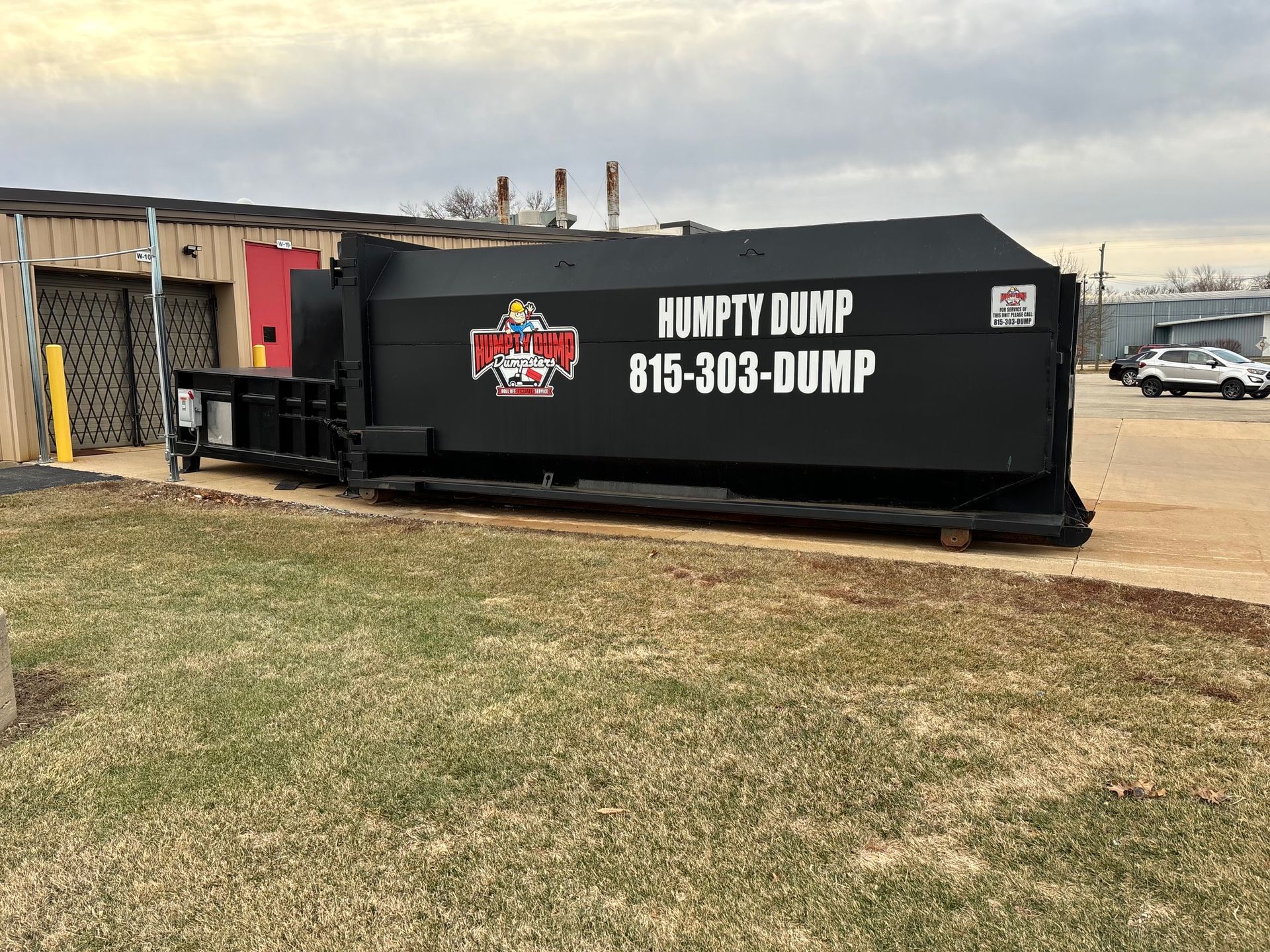 Black Humpty Dump dumpster with phone number 815-303-DUMP, parked near a building and grass.