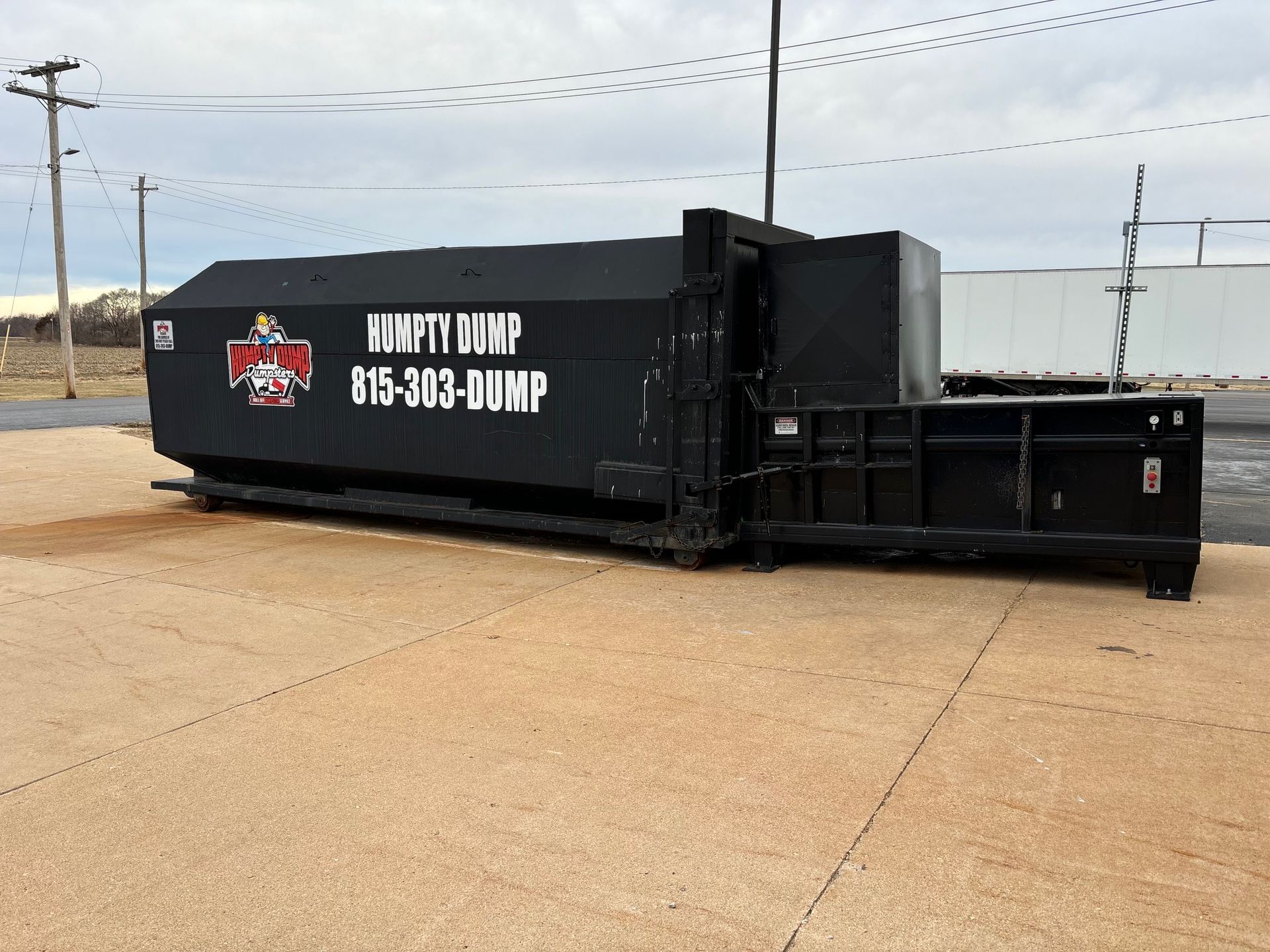Black dumpster with Humpty Dump logo and phone number, sitting outside on concrete, overcast sky.