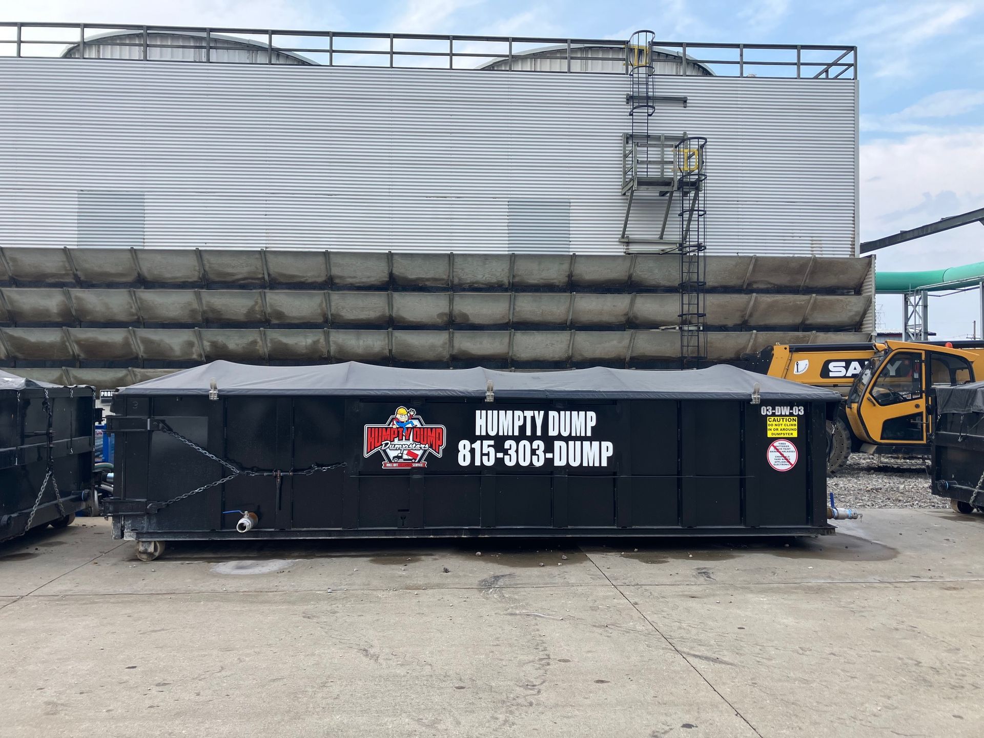 Black dumpster with “Humpty Dump” logo and phone number in front of a factory building.