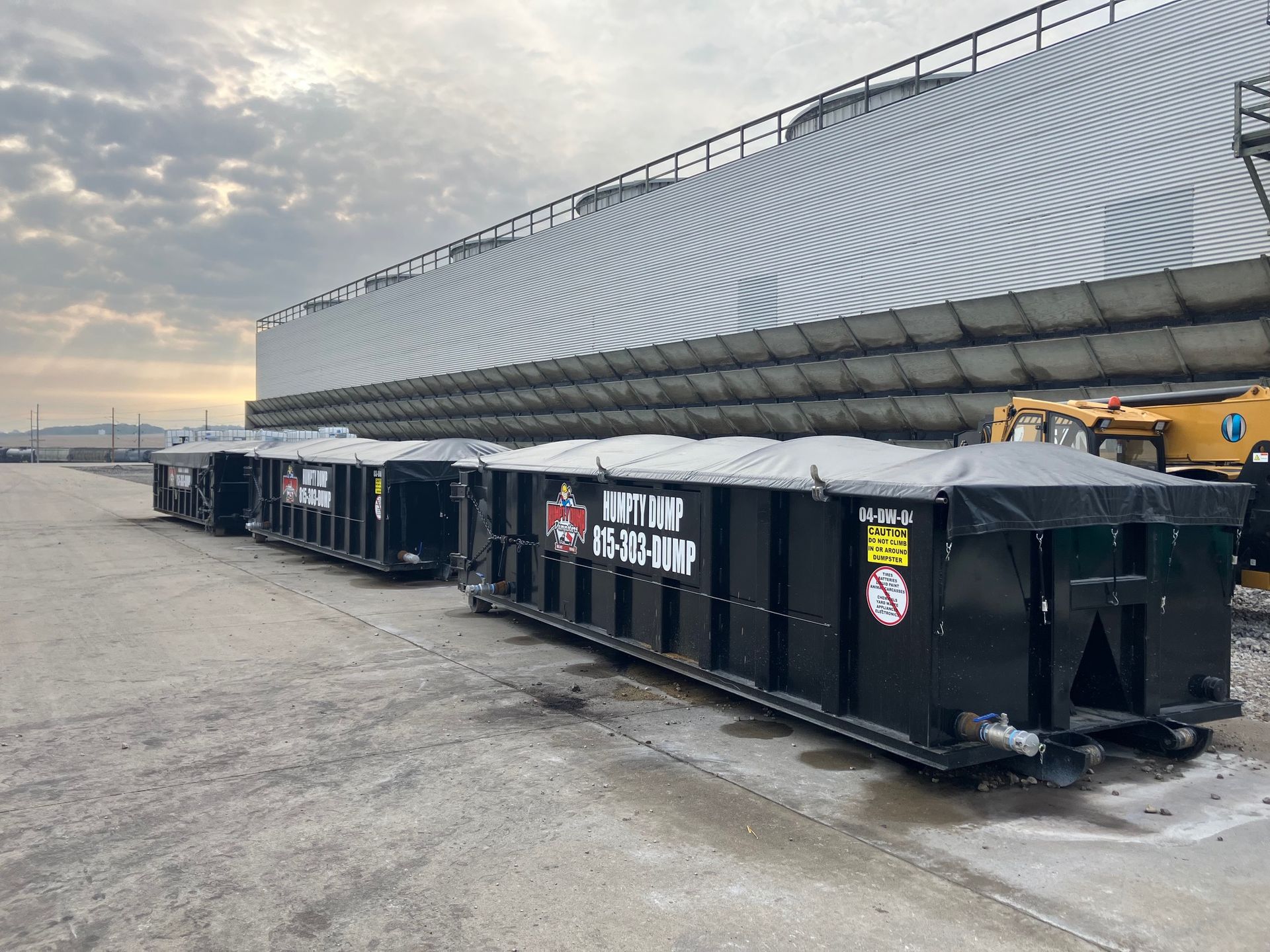 Black roll-off dumpsters lined up on a concrete surface, with a large metallic building in the background.