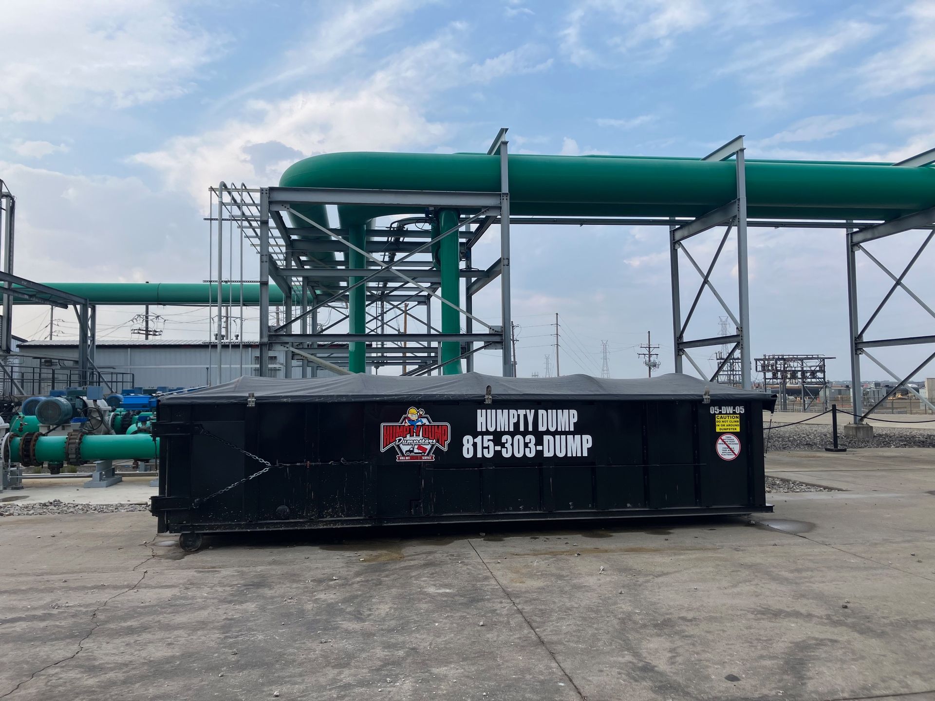 Black dumpster in an industrial setting, green pipes overhead, cloudy sky.