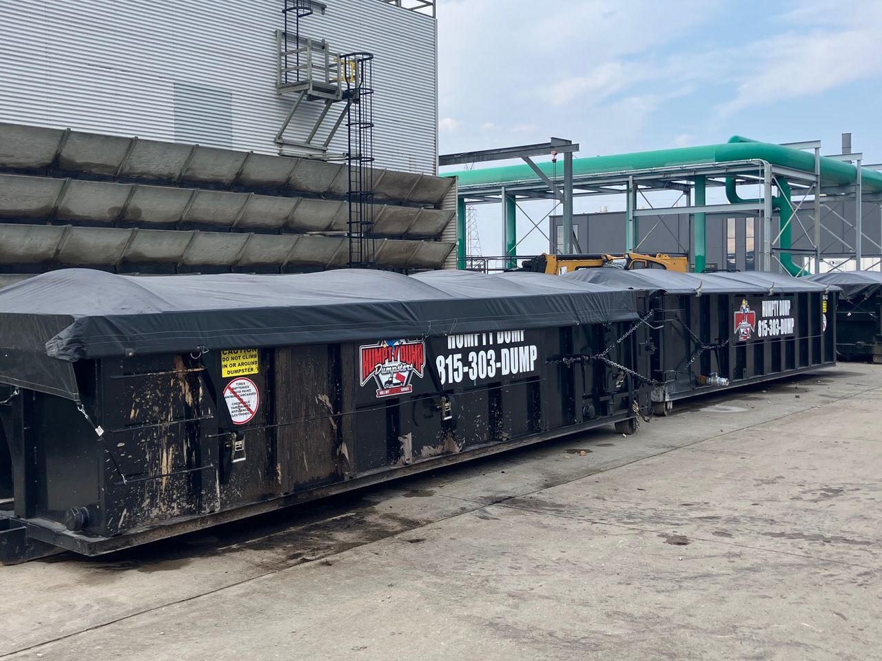 Learn More About Garbage and Rubbish Pickup Black dumpsters in an industrial setting, covered with tarps. A building and pipes in the background.