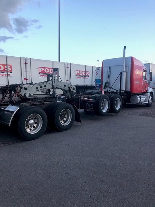 A red and white semi-truck with a specialized flatbed trailer parked in an outdoor lot near white PODS storage containers.