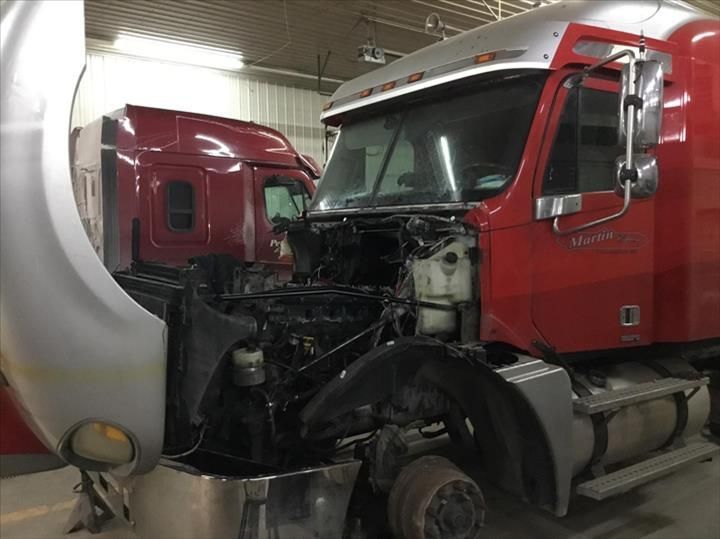 A red semi-truck with its hood open in a garage, revealing the engine compartment, with another truck parked behind it.
