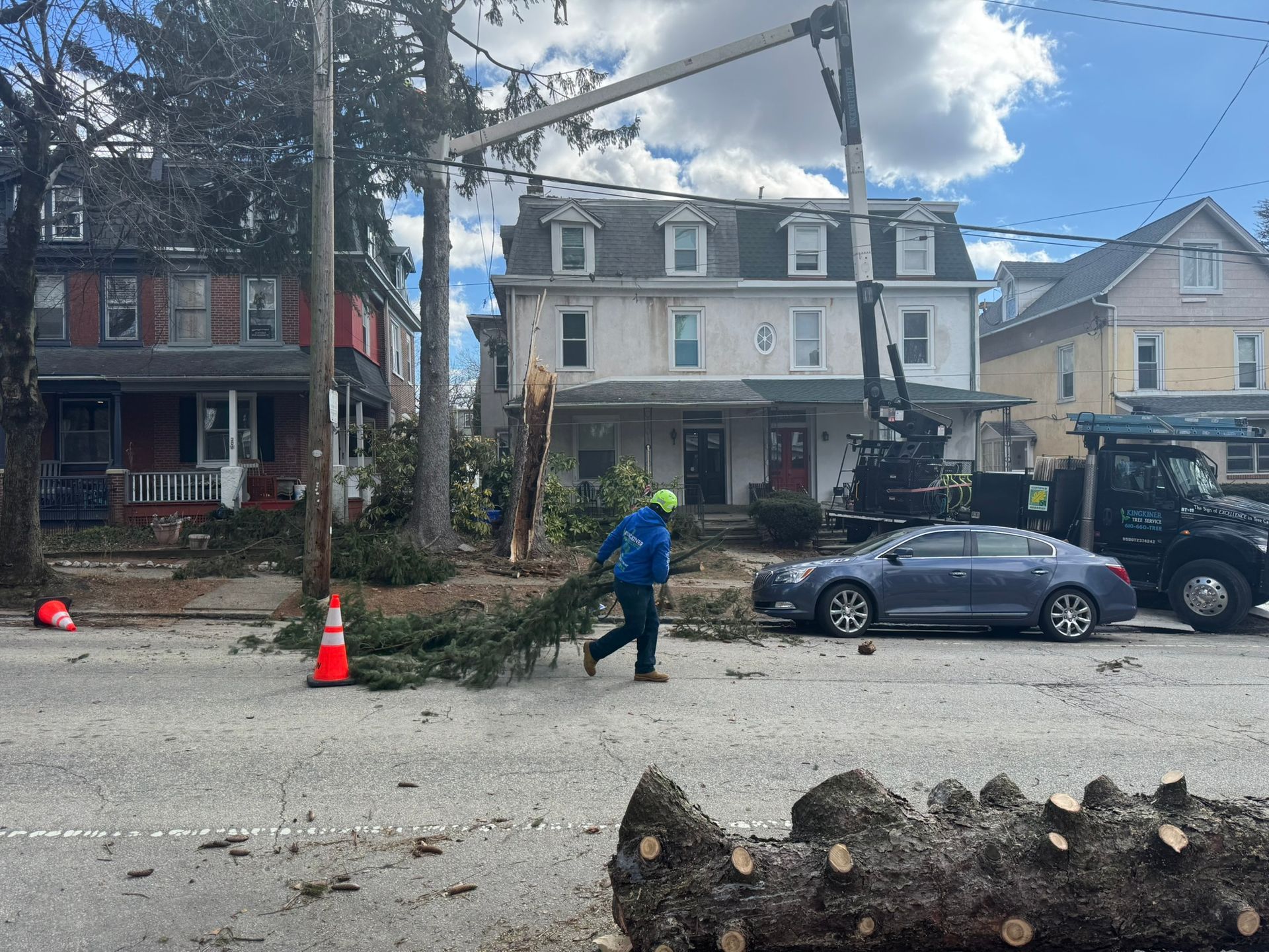 Man carrying tree branch on street, utility truck, damaged house with tree limb removal in progress.