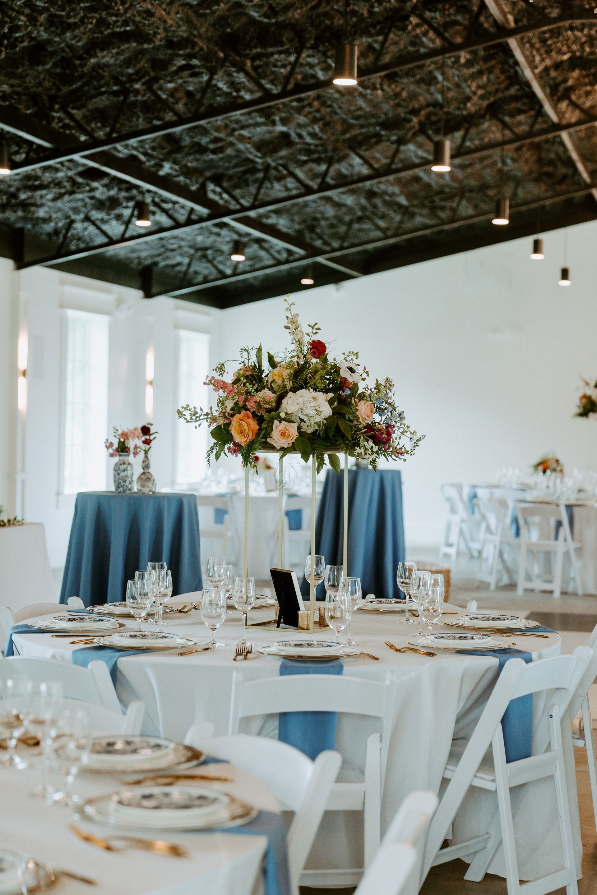 Wedding reception with flower centerpiece on round table, blue tablecloths, white chairs, and venue interior.