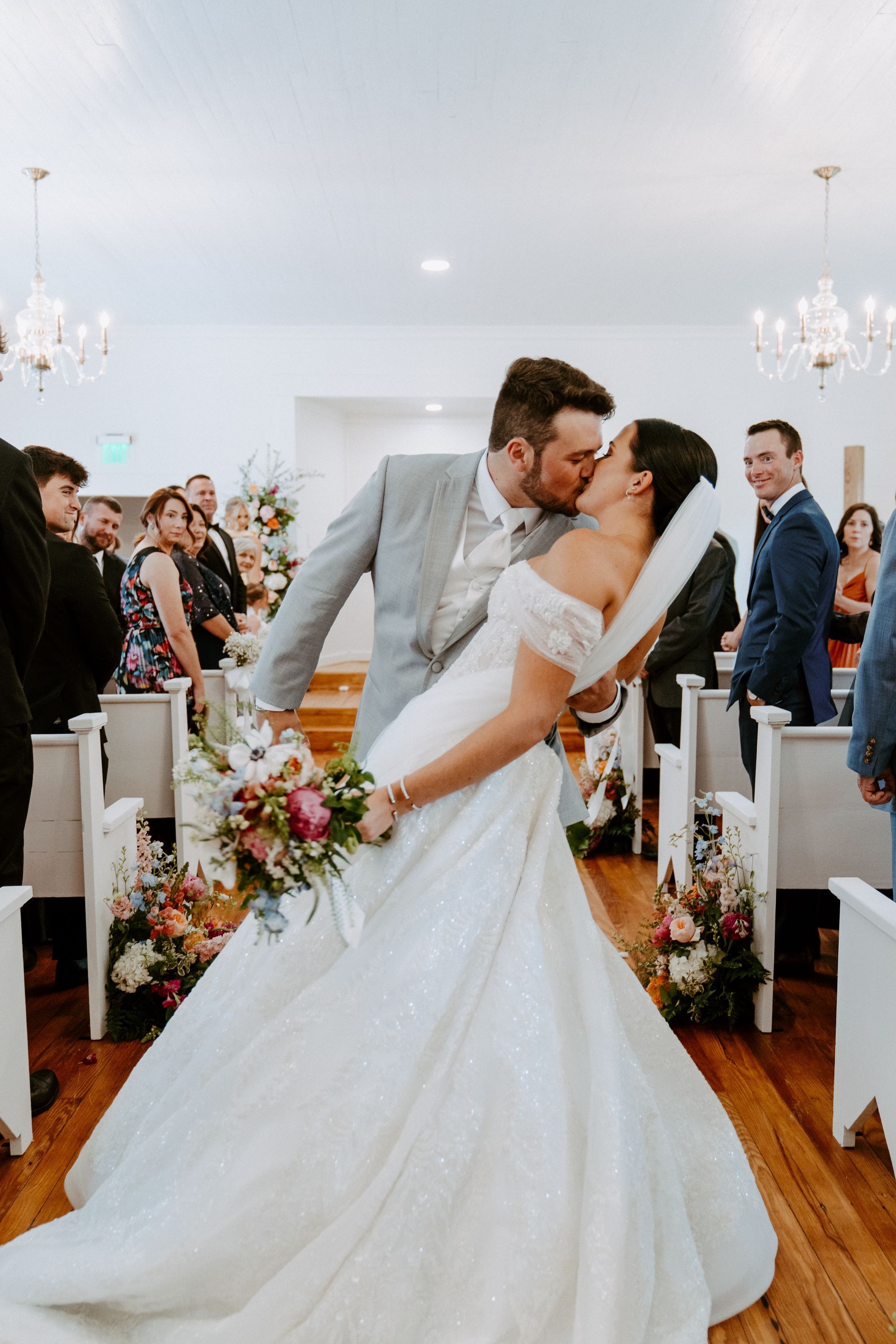 Bride and groom kiss after wedding ceremony. Bride leans back; groom lifts her. Guests cheer. Inside a chapel.
